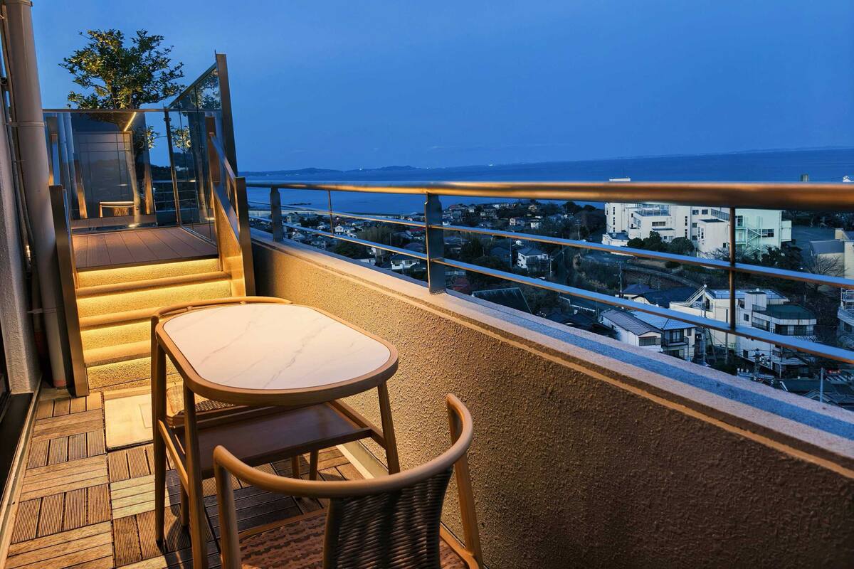 Een balkon met een tafel en stoelen, uitzicht op de stad, en een trap naar een hoger gelegen niveau.