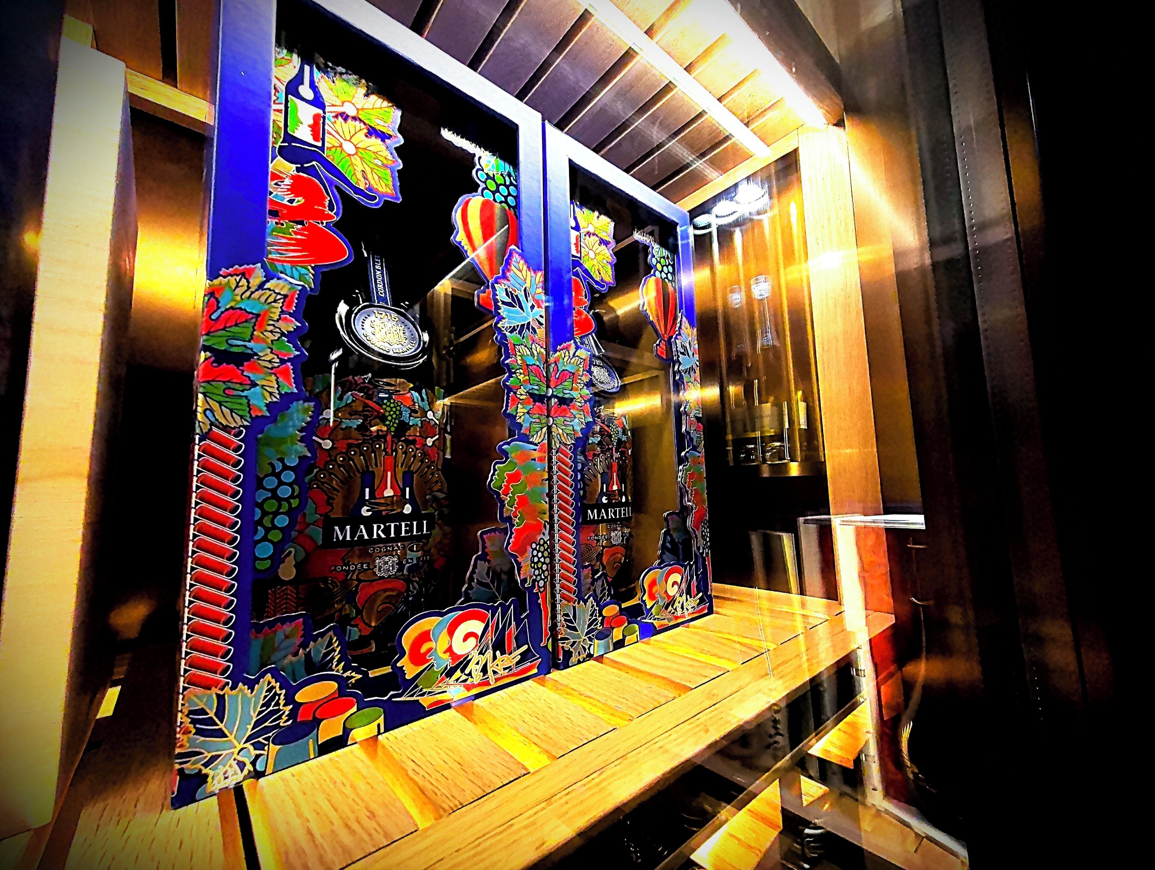 A window display with Martini Cognac bottles, colorful artwork, and a wooden bench.