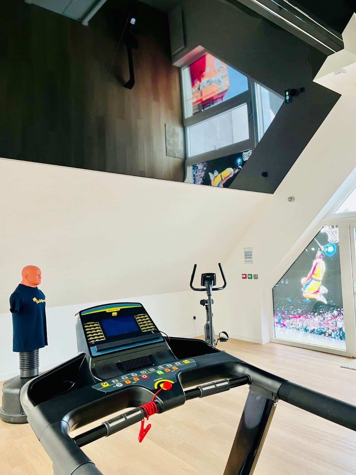 Une salle de sport moderne équipée d’un tapis de course, d’un vélo d’exercice et d’une grande fenêtre donnant sur un immeuble.