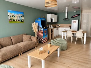 A modern living room with a sofa, a coffee table, a dining area with chairs, a kitchen with cabinets and appliances, and a painting of a cityscape on the wall. - San Francisco - Apartment in a complex with a pool (Neuf-Mesnil)