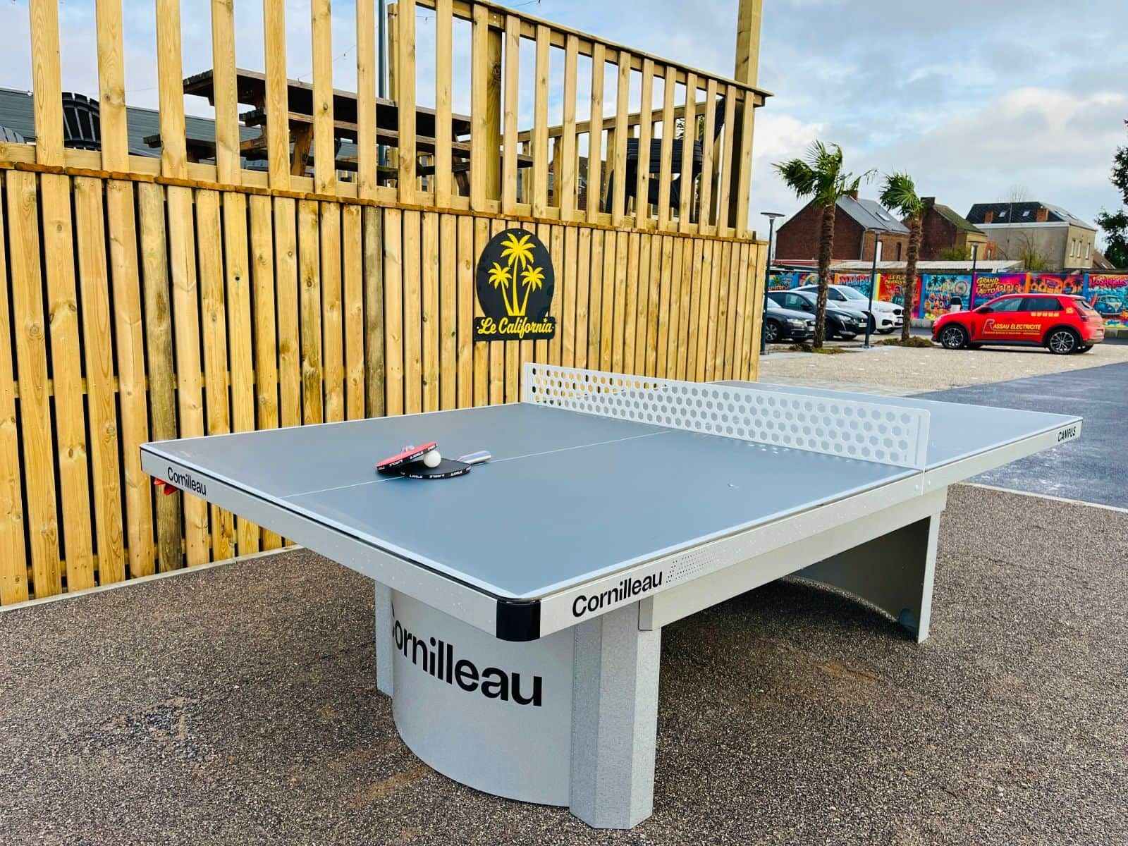 Une table de ping-pong de la marque « Cornilleau », installée à l’extérieur, adossée à une clôture en bois, avec un palmier et un bâtiment en arrière-plan.