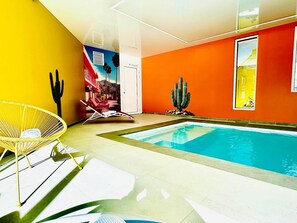 A modern indoor pool area with a yellow and orange color scheme, a pool, lounge chairs, and a cactus decoration.