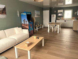 A modern living room with a sofa, a wooden coffee table, a dining area with chairs, a kitchen with cabinets, and a large poster on the wall.