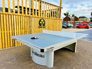 Una mesa de tenis de mesa con la marca 'Cornilleau', ubicada al aire libre junto a una cerca de madera y un edificio con palmeras al fondo.