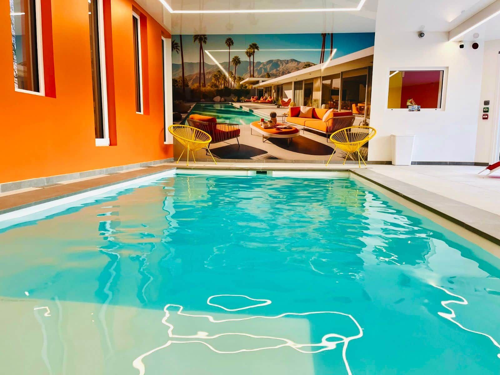 Una piscina con un mural de paisaje tropical, paredes anaranjadas y sillas amarillas.