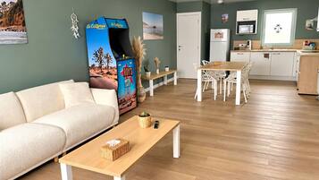 A modern living room with a sofa, a coffee table, and a dining area with chairs. There is a kitchen area with a refrigerator and a microwave.