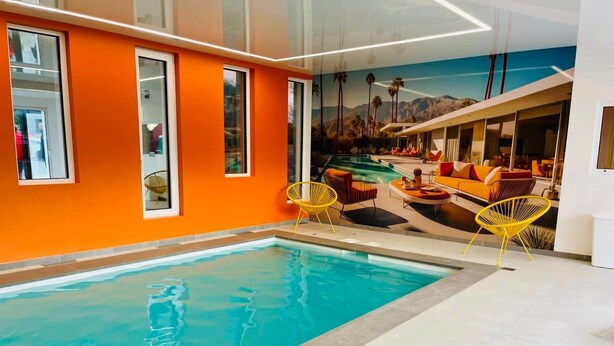 Una piscina interior con un mural de paisaje tropical, paredes color naranja y sillas amarillas.
