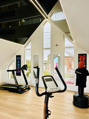 Une salle de sport moderne équipée de deux tapis de course, d’un vélo elliptique et d’un vélo d’appartement. De grandes fenêtres laissent entrer la lumière naturelle et offrent une vue sur l’extérieur.