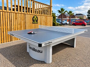 Une table de ping-pong de la marque « Cornilleau », installée à l’extérieur, avec une clôture en bois et un palmier en arrière-plan.
