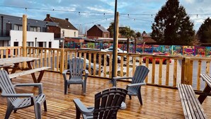 Una terraza de madera con sillas y mesas de plástico, un poste de metal y una cerca adornada con colores.