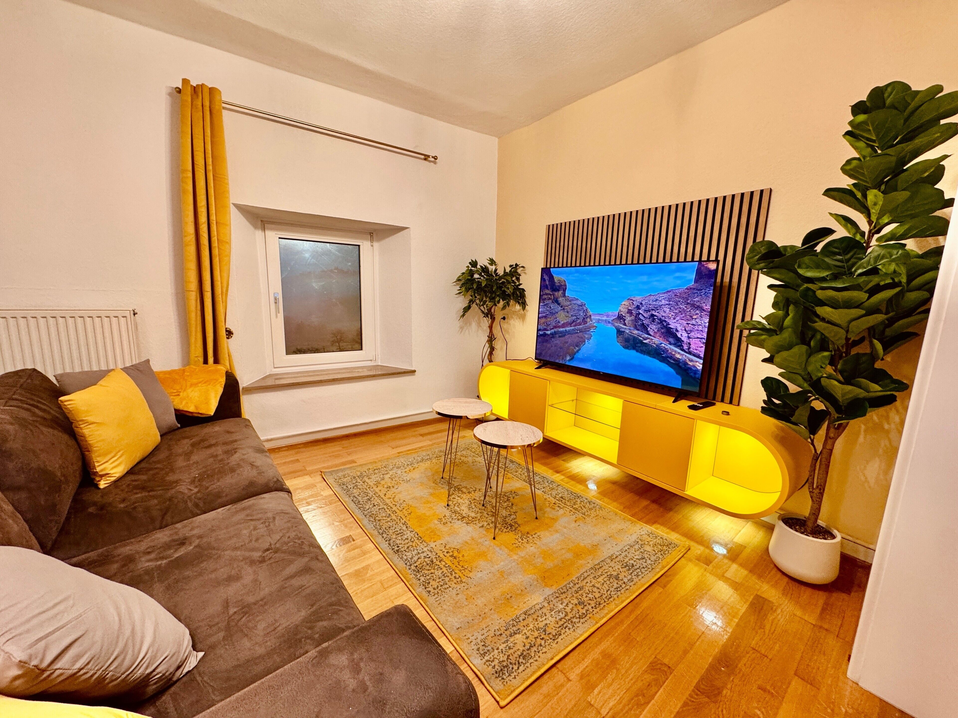 Ein Wohnzimmer mit einer Couch, einem Fernseher, einem Fenster, einer Pflanze und einem gelben Schrank.