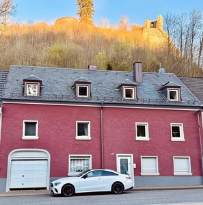 A red building with a white car parked in front, a garage, and a hill in the background.