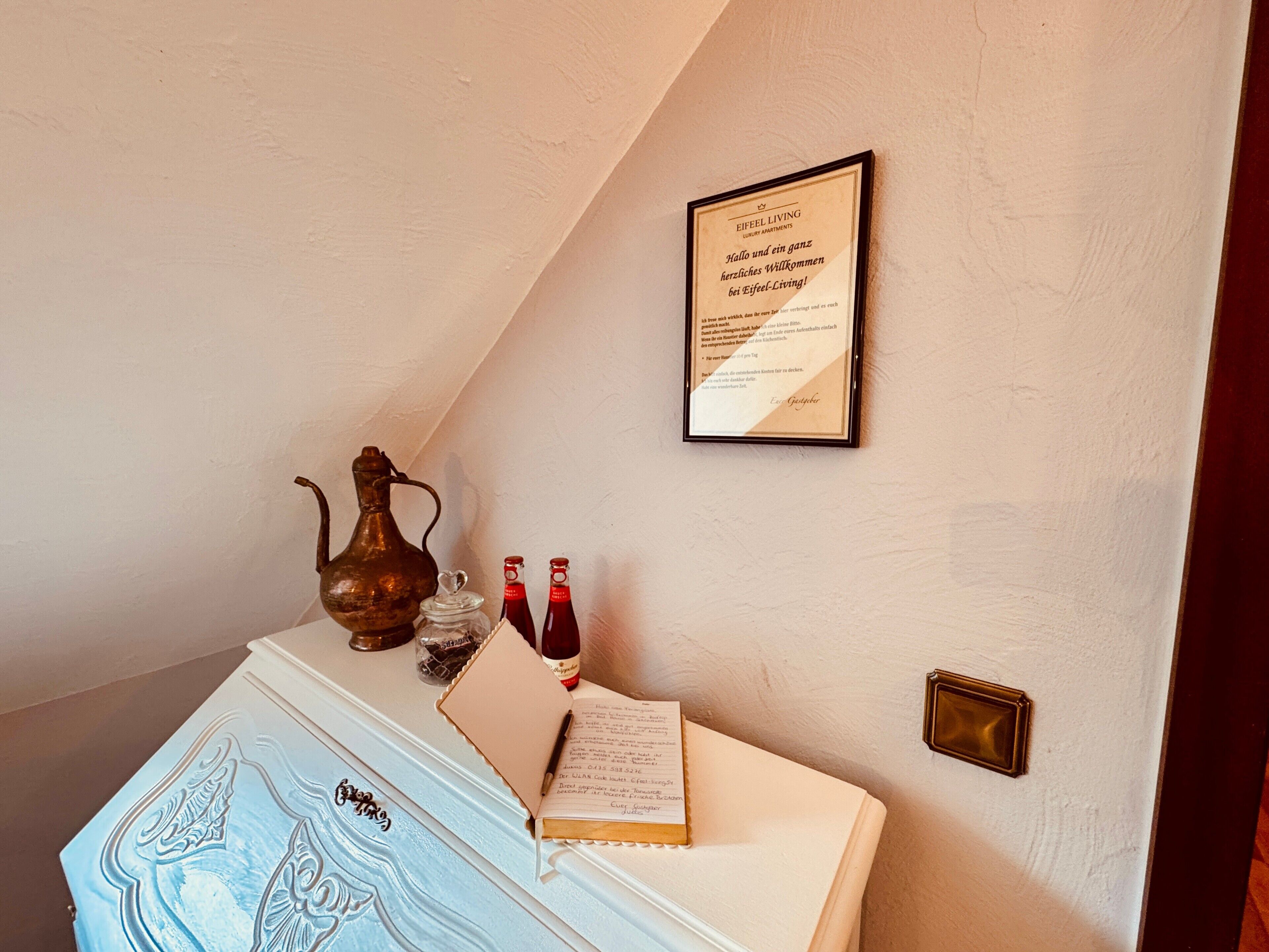 Ein Zimmer mit weißer Wand, einem gerahmten Willkommensschild, einer Vintage-Vase und einem blauen Schrank mit dekorativem Muster.
