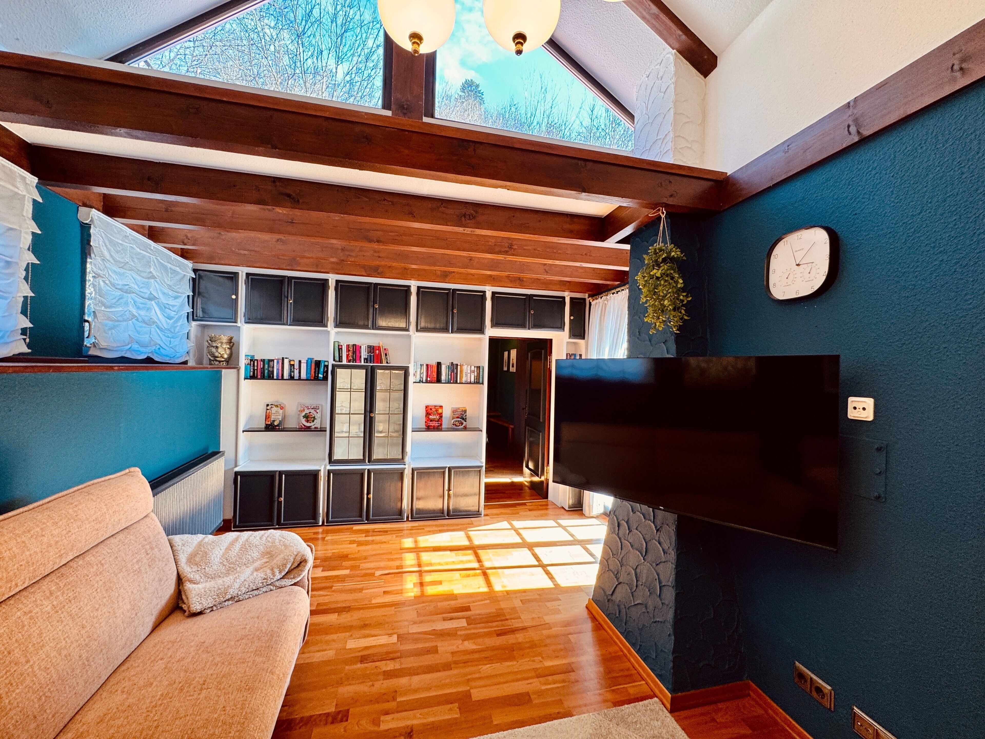 Ein Wohnzimmer mit Holzboden, einer Couch, einem an der Wand montierten Fernseher und einem Bücherregal voller Bücher.