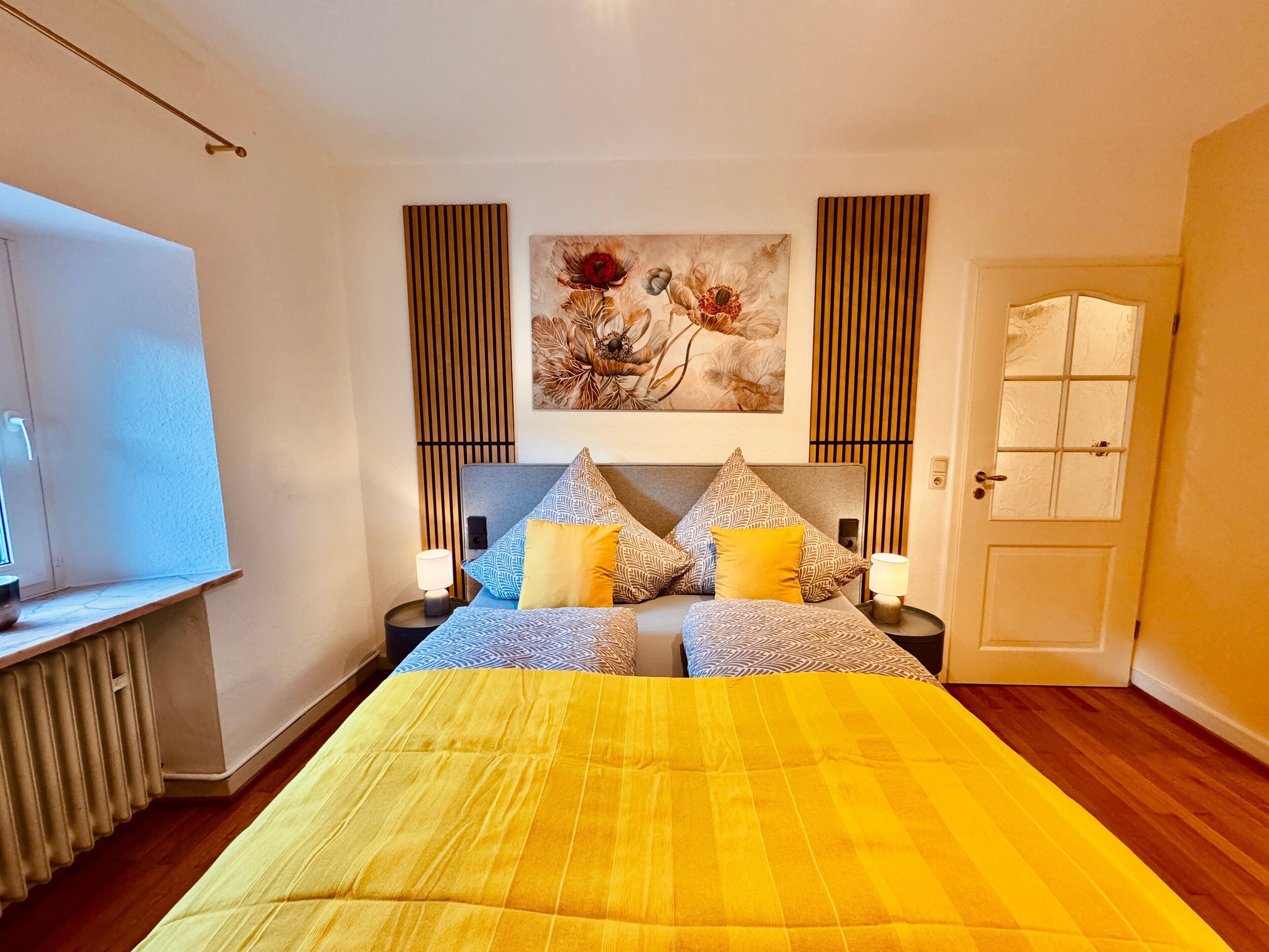 A bedroom with a bed, two bedside tables, a radiator, a door, and a painting on the wall.