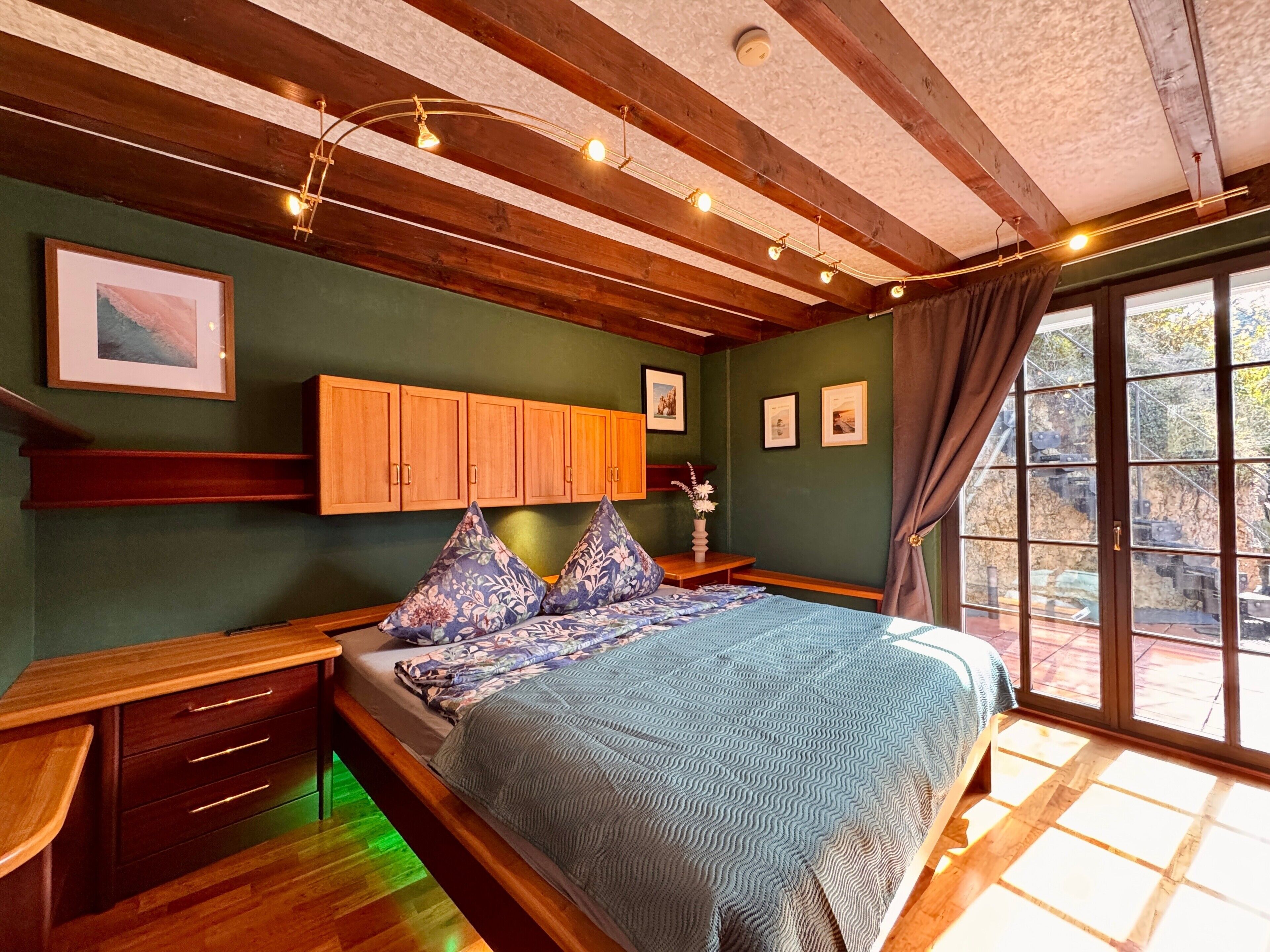 Une chambre à coucher avec un lit, une tête de lit en bois, une armoire intégrée et de grandes fenêtres avec des rideaux.