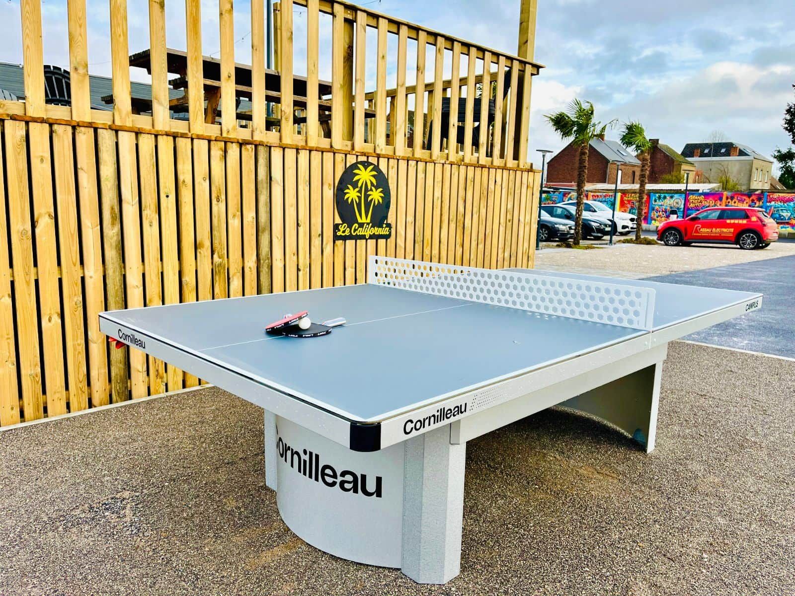 Une table de ping-pong de la marque « Cornilleau », placée devant une clôture en bois, avec un palmier et un bâtiment en arrière-plan.