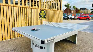 Une table de ping-pong de la marque « Cornilleau », placée devant une clôture en bois, avec un palmier et un bâtiment en arrière-plan.