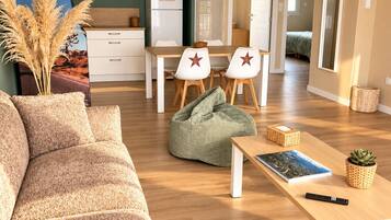 Un salon moderne avec un canapé, une table à manger et des chaises, un fauteuil poire, une petite table avec un livre et une télécommande, et un coin cuisine à l’arrière-plan.