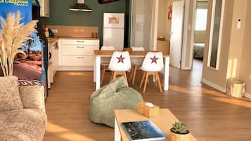 Een moderne woonkamer met een bank, een bijzettafeltje met een boek en afstandsbediening, een groene relaxstoel en een eethoek met stoelen en een tafel.