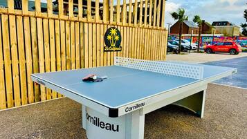 A ping pong table with 'Cornilleau' branding, set outdoors with a wooden fence and palm trees in the background.