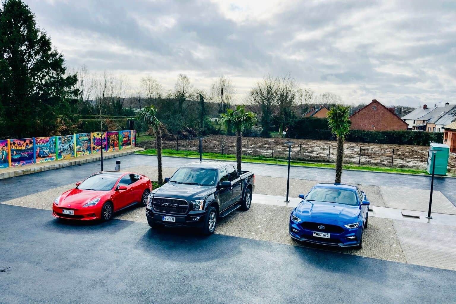 Trois voitures sont garées sur un parking. On aperçoit une fresque murale sur le côté gauche et un coffret électrique vert sur le côté droit.