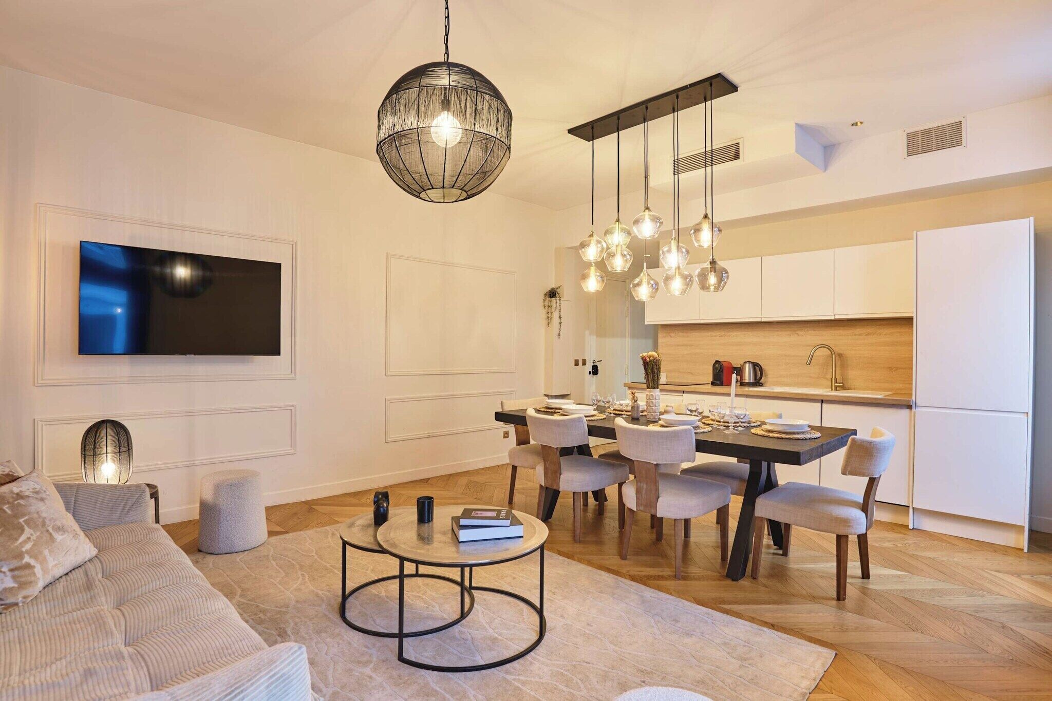 A modern living room with a sofa, a dining table, and chairs, a kitchen area with cabinets and a countertop, and a large flat-screen TV mounted on the wall.