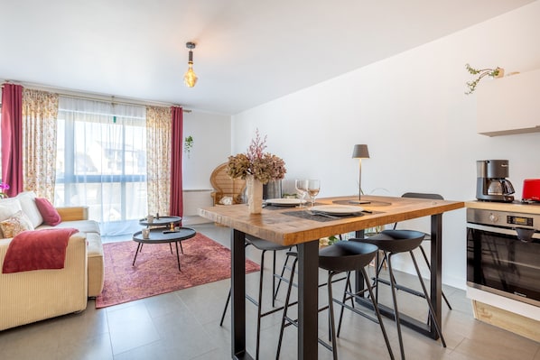 A modern living room with a sofa, a dining table with chairs, a coffee machine, and a kitchen area.