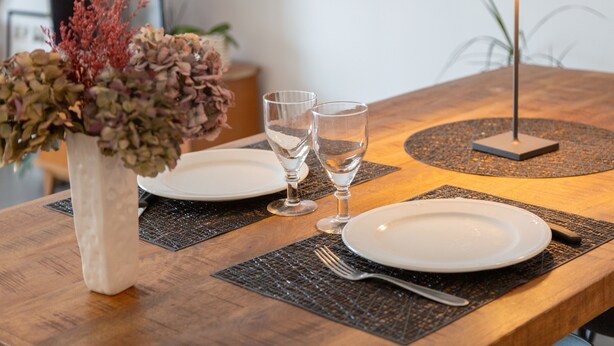 A dining table with two plates, two glasses, a vase with dried flowers, a lamp, and a placemat. - Ty Elise – Charming Central Apartment in the Heart of Quimper (Quimper)