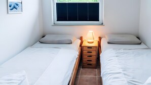 A small, clean room with two single beds, a nightstand with a lamp, and a window with blinds.