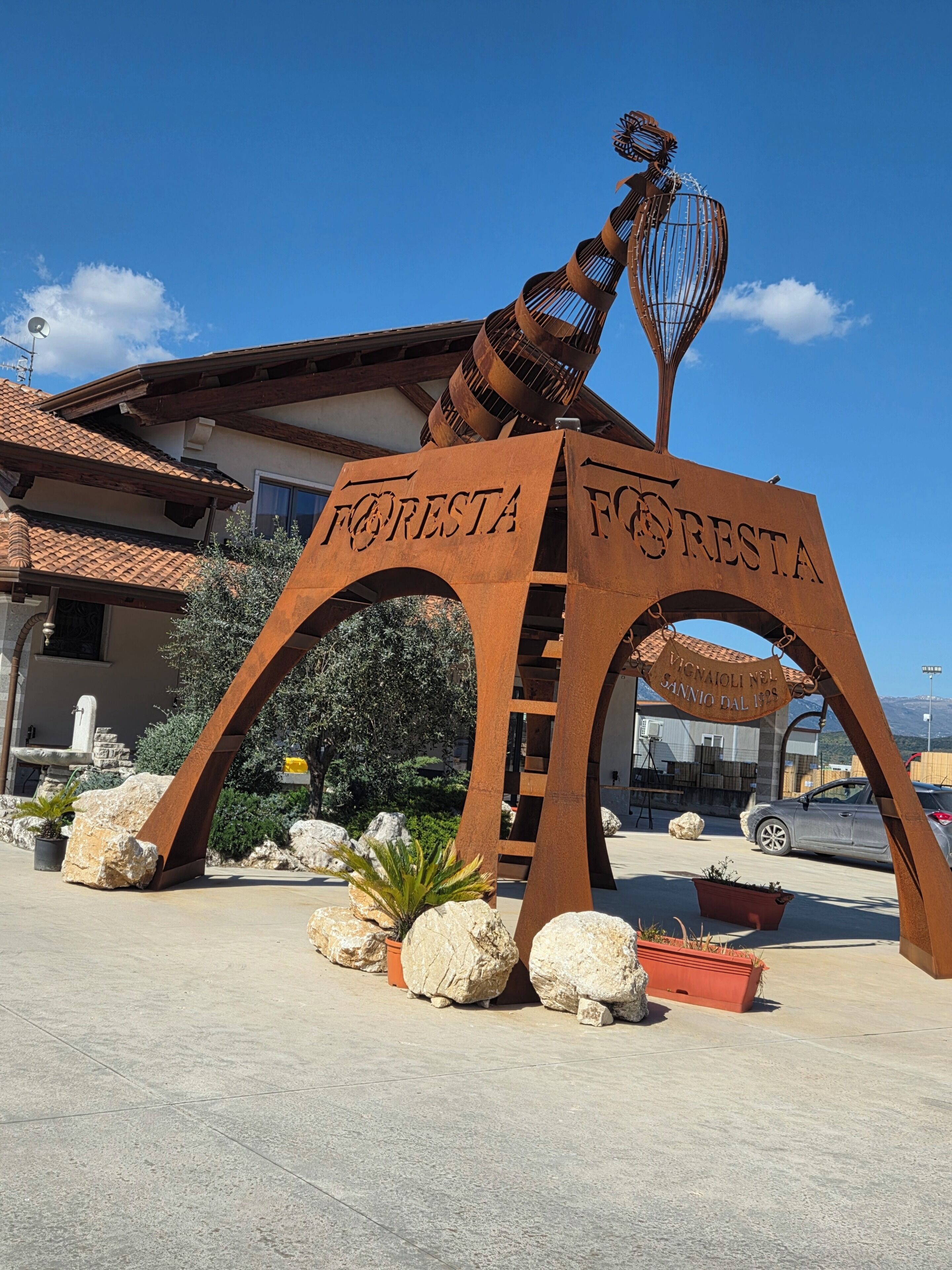 Un arco de metal con 'FORESTA' escrito, que da paso a un edificio de tejado a la vista.