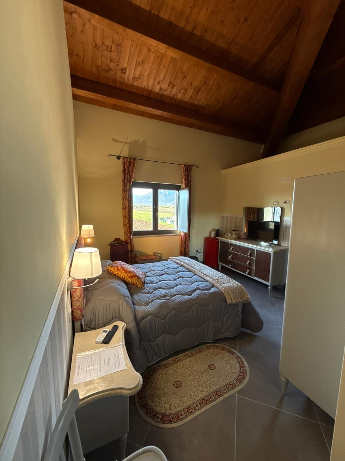 A bedroom with a bed, bedside table, lamp, and a view of the outside.