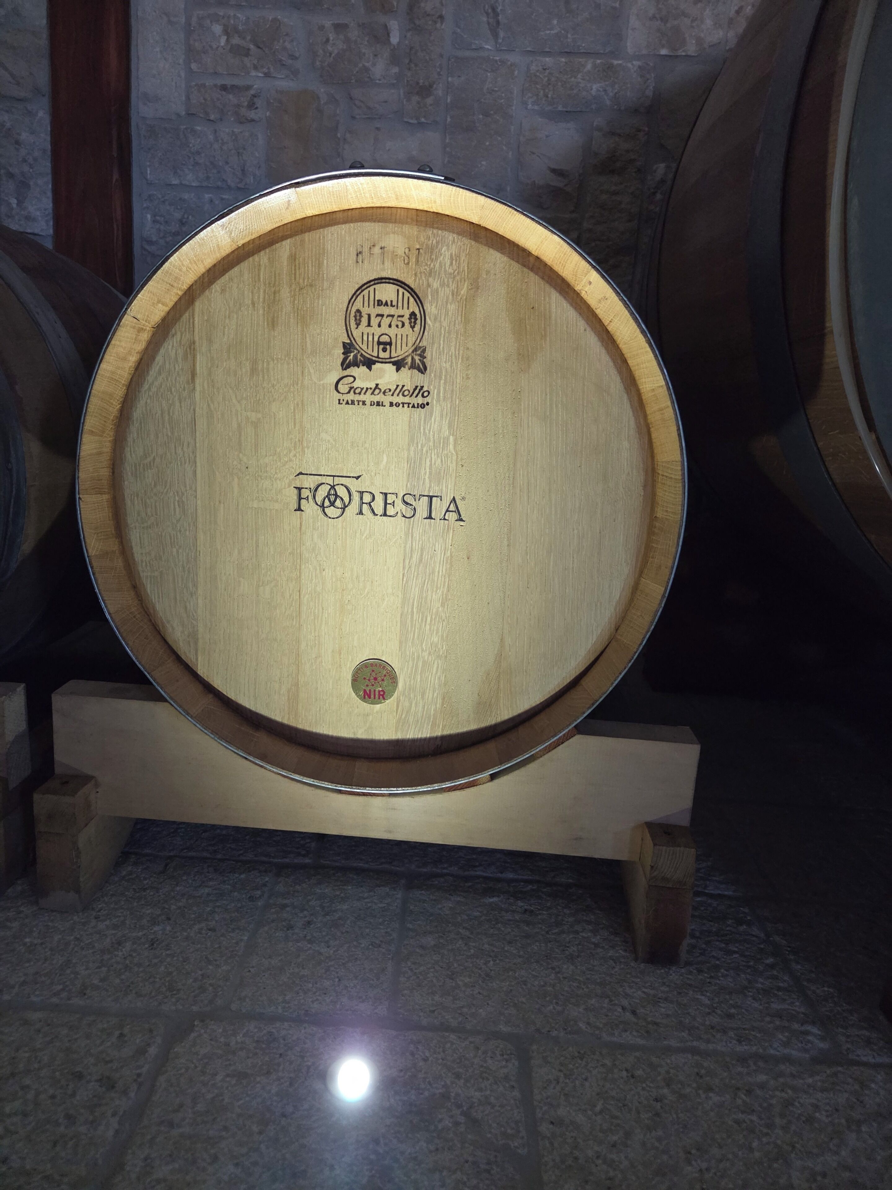 A wooden barrel with 'FORESTA' and 'NIR' labels, placed on a stand in a dimly lit room with stone walls.