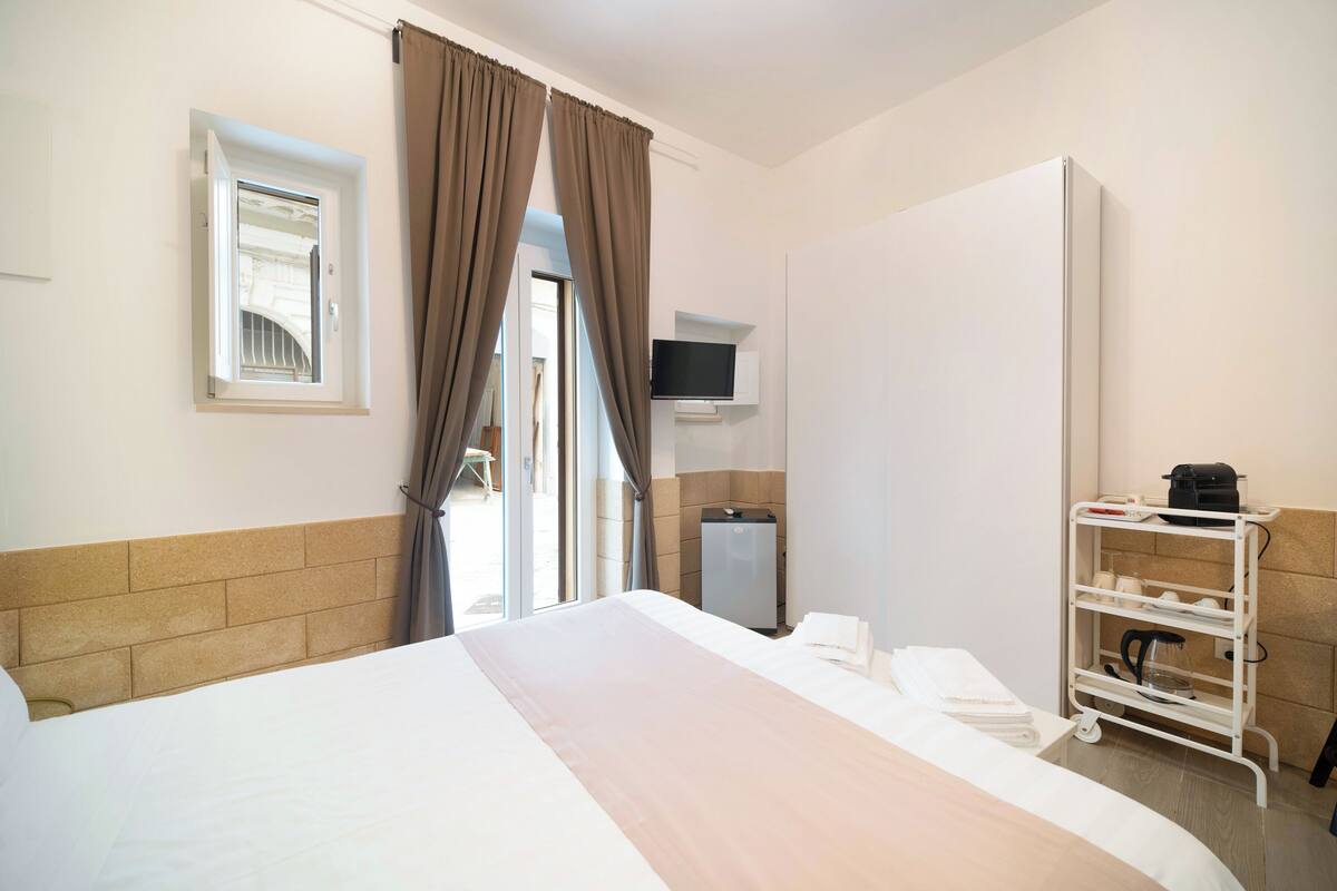 A hotel room with a bed, a TV mounted on the wall, a wardrobe, a small desk with a kettle, and a window with curtains.