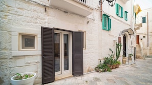 A narrow street with a building featuring a door with shutters, a potted plant, and a cactus. - Le Contrade Vico Alba (Monopoli)