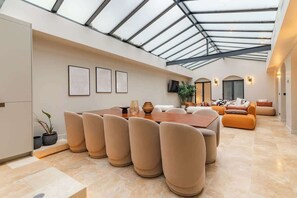 Une pièce spacieuse avec un plafond haut et un puits de lumière, comprenant un coin repas avec une table et des chaises en bois, un ensemble de canapés et un téléviseur fixé au mur.