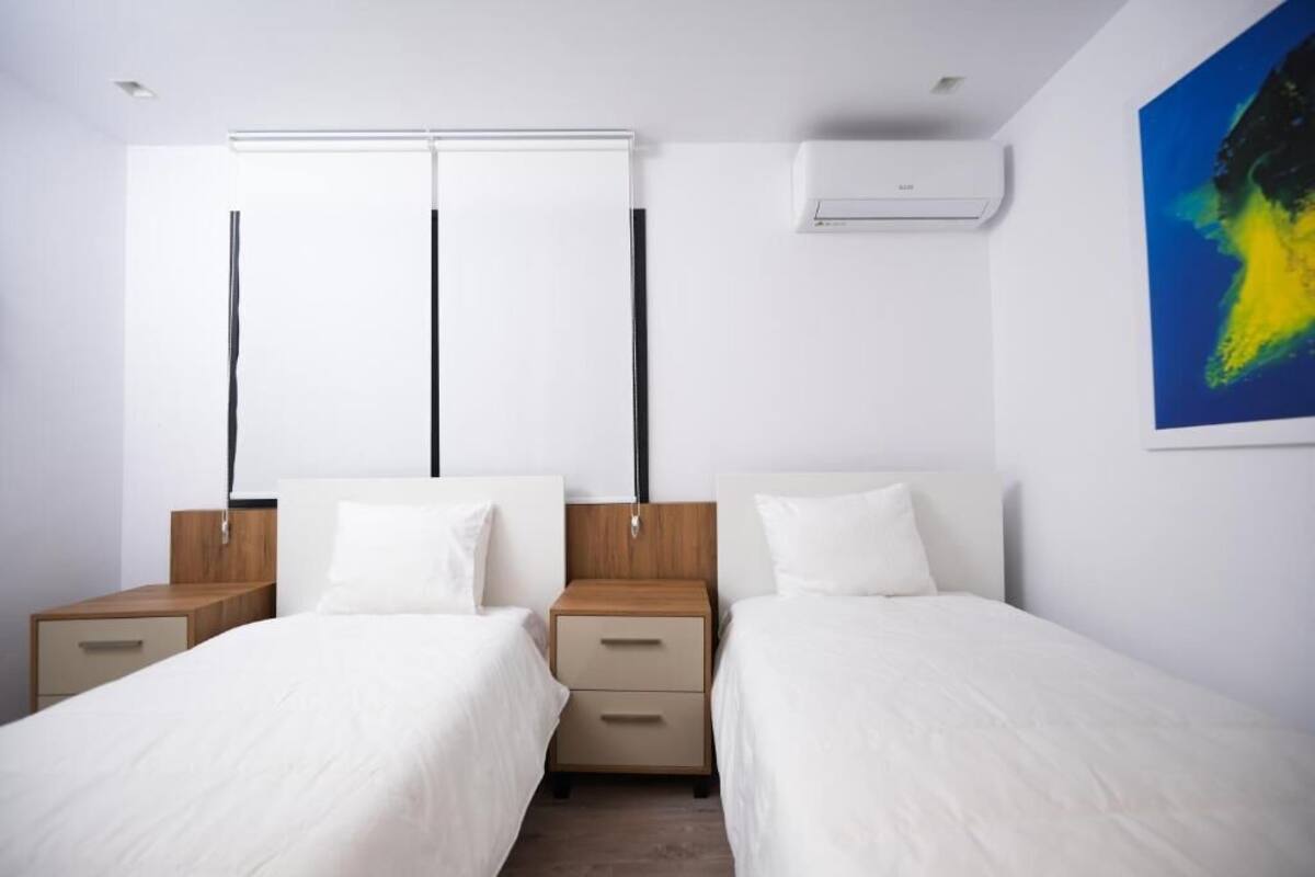 A modern hotel room with two beds, a nightstand with drawers, a wall-mounted air conditioner, and a large abstract painting.