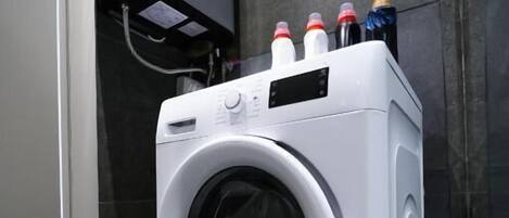 Une machine à laver moderne et compacte, installée dans une salle de bain aux carreaux foncés, avec un chauffe-eau apparent.