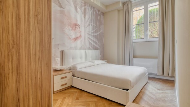 Une chambre à coucher comprenant un lit, une table de chevet, une œuvre d’art accrochée au mur et une fenêtre avec des rideaux.