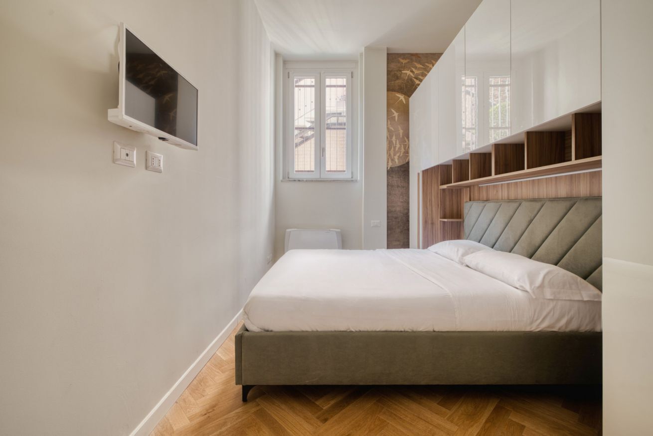 A bedroom with a bed, built-in shelving, a TV, and a window.