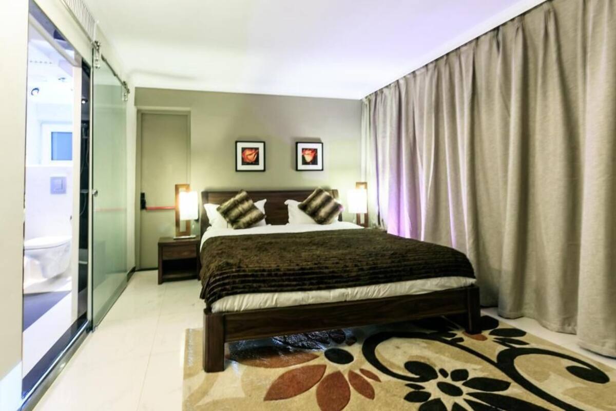 Una habitación de hotel con cama, mesita de noche, lámpara, ventana con cortinas y puerta.