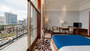 A hotel room with a balcony, a bed, a desk with two chairs, a television, and a view of the cityscape.