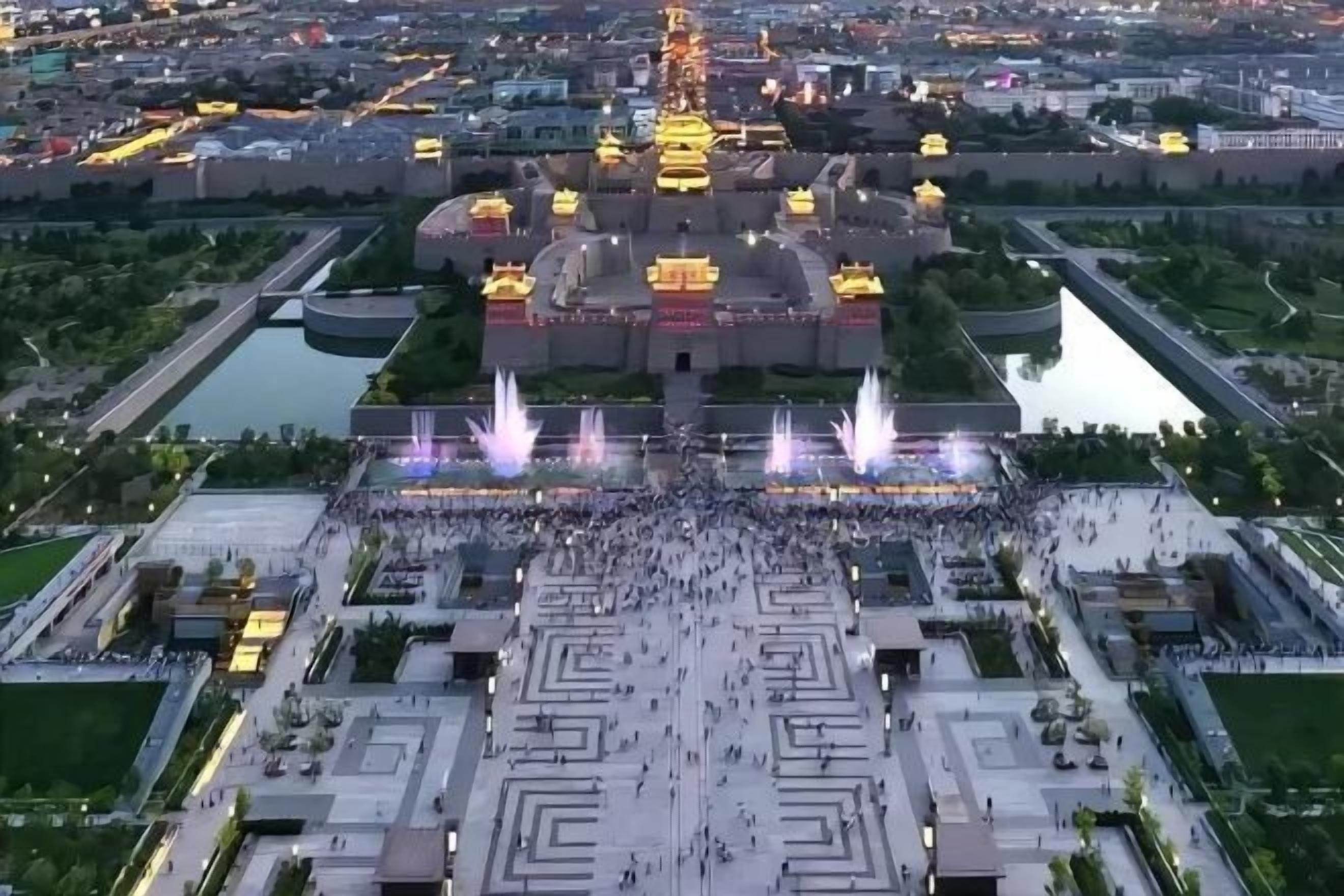 An aerial view of a large public square with a central monument, surrounded by greenery and water features.