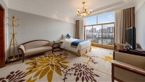 A hotel room with a bed, a sofa, a TV, a large window with a city view, and a chandelier.