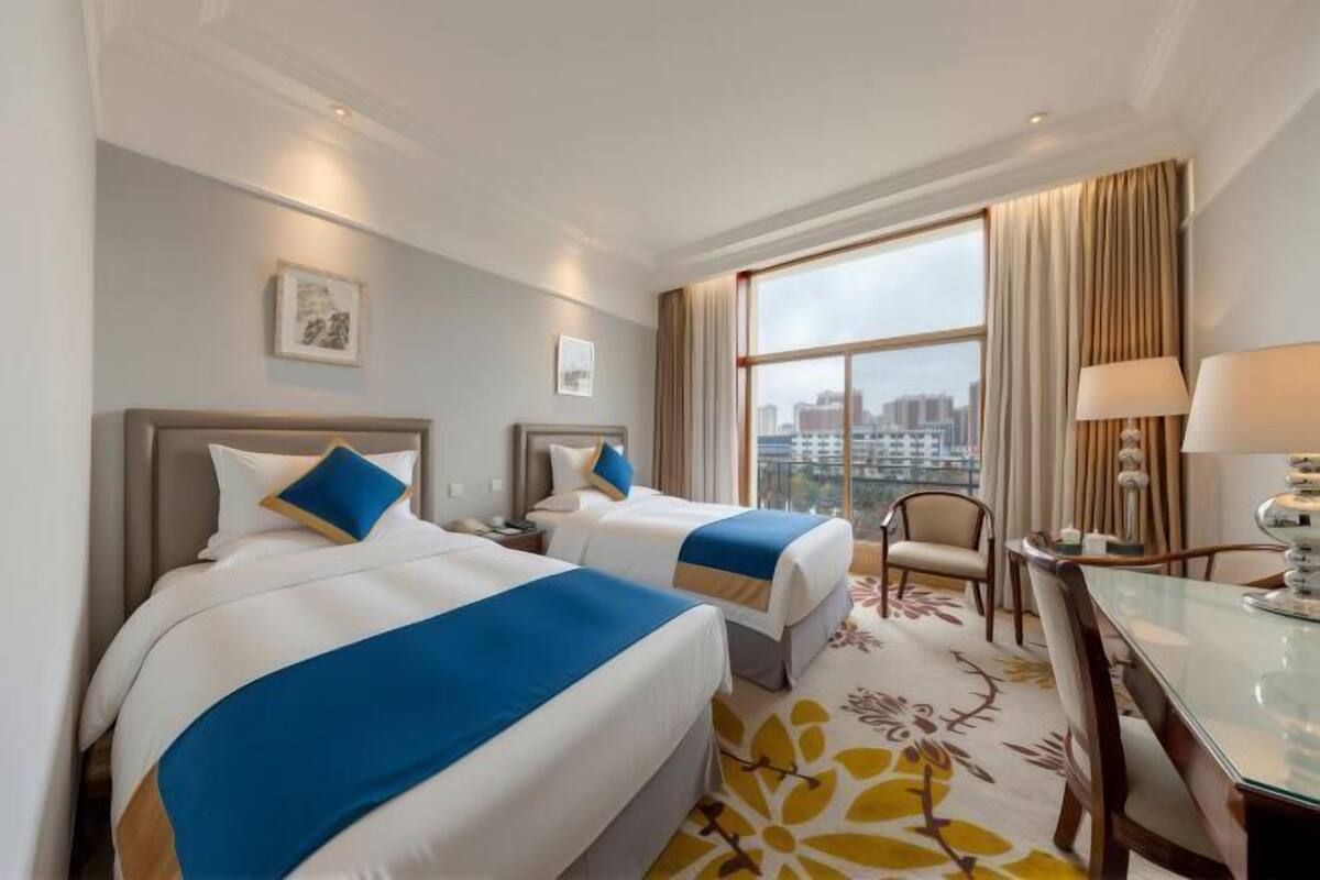 A hotel room with two beds, a desk, and a large window with a city view.