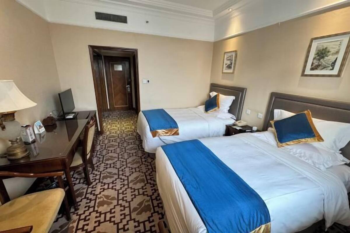 A hotel room with two beds, a desk with a lamp, a chair, and a patterned carpet.