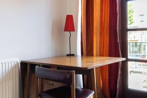 A corner room with a wooden table, a red lamp, a chair, and a window with curtains.