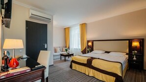 A hotel room with a bed, a desk with a lamp, a chair, a small table, and a window with curtains.