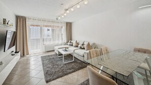 A modern living room with a sofa, a glass coffee table, a dining table, and a TV.