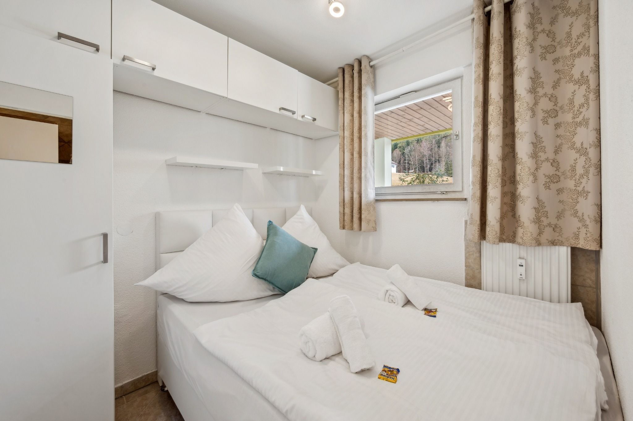 Een netjes opgemaakt bed met witte lakens, een blauwgroen kussen en een handdoek, een witte garderobe met spiegel en een raam met gordijnen.
